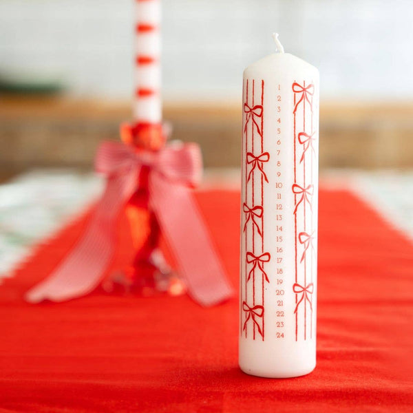 White candle with red bow design on a red surface, with blurred decorative items in the background.