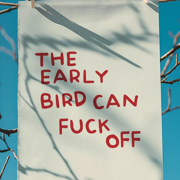 White towel with red text 'THE EARLY BIRD CAN F*** OFF' against a blue sky with tree branches.