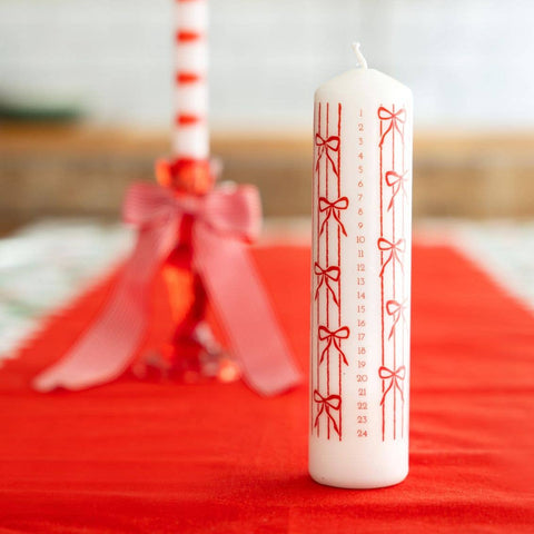White candle with red bow design on a red surface, with blurred decorative items in the background.