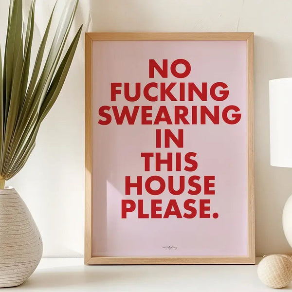 Framed poster with red text 'NO FUCKING SWEARING IN THIS HOUSE PLEASE.' on a pink background, placed on a white surface with decorative elements.