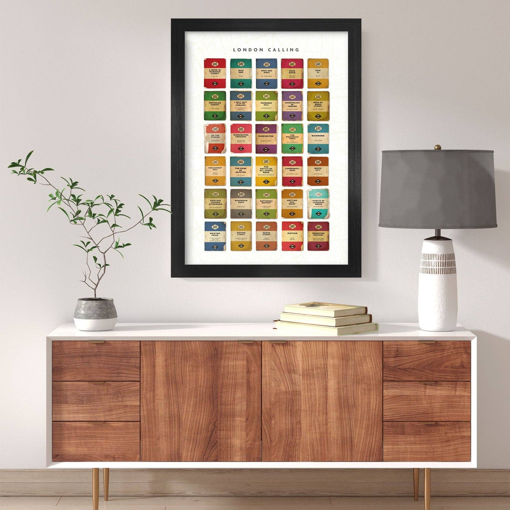 Framed art print on a wall above a wooden sideboard with books and a lamp.